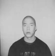 Black and white photograph of Wayne Lo. Shaved head in a black t shirt standing against a white brick wall.