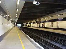 Several people walking inside a building with a black ceiling and white walls bordered in black and displaying several advertisements