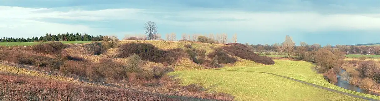 At left a grassyu hill with shrubbery, dropping to a meadow at centre. At right a narrow river winds by.