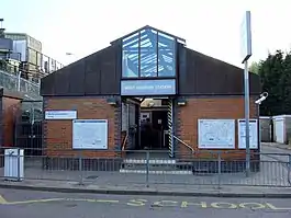 A brown-bricked building with a rectangular, light blue sign reading "WEST HARROW STATION" in white letters all under a light blue sky