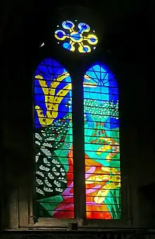 A stained-glass window in bright primary colours depicting a path in the countryside with trees and plants on either side.