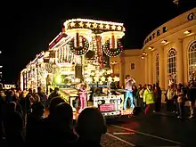 A large vehicle lit by multi-coloured lamps and carrying people in brightly coloured coustumes in front of a large crowd on a darkened street.