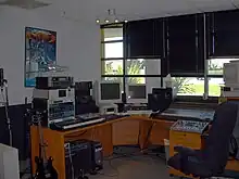 A musician's office with a solid wall and a wall containing windows shown, a microphone stand, an electric guitar, a mixing board, a keyboard, two computer monitors and a computer, a poster of the Transformers franchise