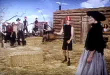 A  man wearing a black turtleneck and a red hat holds a whip while a woman in a black dress smoking a cigarette with a holder stands in front of him. In the background are onlooking cowboys and cowgirls, four men wearing black turtlenecks playing instruments, and a ranch house.