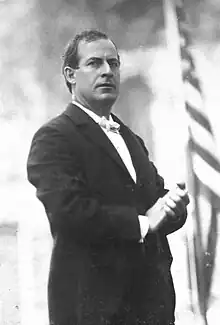 A man in his thirties wearing a dark suit holds his hands together in front of him as he looks to his right. In the background can be seen the stars and stripes.