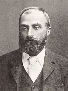 Heavily bearded man with dark receding hair, wearing a dark coloured jacket, white collar and pale tie. He is looking slightly to the left, with a solemn expression