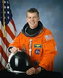 man in space suit holding helmet, American flag in background