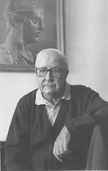 An older white man in glasses, facing the camera, sitting in front of a picture of the Delphi Charioteer.