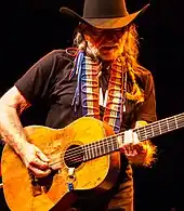 A man with long white hair and white beard playing a guitar. He wears a black T-shirt, which is crossed by the red, white and blue strap of the guitar. He also wears black pants.