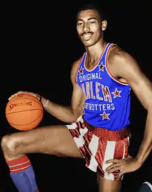 Black man kneeling on one knee with a basketball resting on the other and his hand atop the basketball. He is wearing a red, white and blue Harlem Globetrotters uniform.