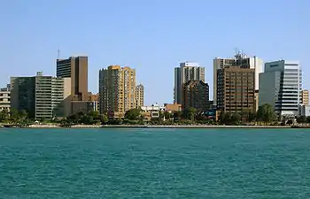 A landscape photograph of a modern North American city beside a river, taken from the other side of the water at a distance of perhaps 300 metres. A number of high-rise buildings are visible as well as a paved promenade on the waterfront.