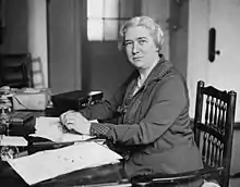 Monochrome photograph of Cullis sitting at her desk and looking over her left shoulder to the camera. There is a parcel and suitcase on the desk.