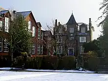 A colour photograph of an unusual Victorian house with a small spire on the top. To the left is a set of old fashioned schoolrooms with large sash windows. In front of the house is a small lawn, covered in snow.