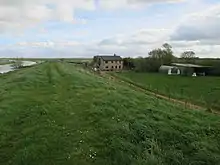 Colour photograph showing a small river on the left, and grass with a cottage on the right