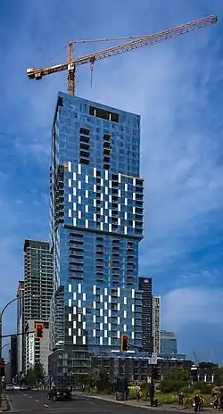 A tall bluish glass-sheathed rectilinear tower with a tower crane atop, rising from an area behind a vacant lot, in front of which is an intersection with red traffic lights. A blue sky speckled with small clouds is in the background.