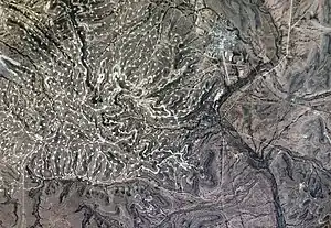 A photograph looking down on hilly, treeless terrain with a town to the upper right and the thin line of a river crossing up and down on the right