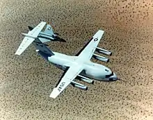 Top view of cargo aircraft in-flight, trailed by a fighter chase aircraft. Under each un-swept wing are two engines suspended forward ahead the leading edge.