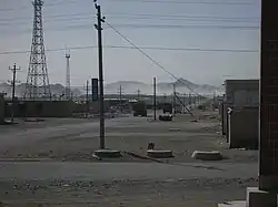 A dusty street with one-storey buildings on each side, a tall radio antenna, lorries on the road and grey mountains in the background.