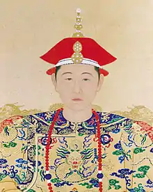 Full-face-view color painting of the head and shoulders of a young man wearing a two-tiered red cap with a white edge, a necklace made of red beads except for two larger blue beads each surrounded by two white beads, and wearing a yellow robe covered with dragon-and-clouds patterns in green, blue, and red.