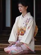 A young woman kneeling in an offwhite formal kimono with a traditionally-stylized pink blossom pattern.