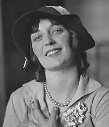 A smiling young white woman, wearing a hat, seated in a chair with one hand against her chest and the other in her lap