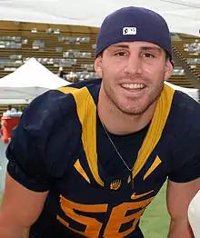 American football player standing with arms akimbo, in gold jersey (#56) and dark blue pants, wearing a dark blue baseball cap