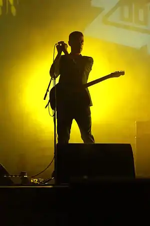 Johnny Blake stands on a stage holding a microphone in both hands. He wears a guitar, there are speakers in front of him and behind him, and a bright green light shines behind him