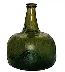 A squat semi-transparent green blown glass bottle, shaped vaguely like a squarish mandarin citrus, with a strongly concave base and a short flanged neck.