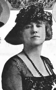 A white woman wearing a dark hat with an asymmetrical brim, and a dark dress with a square neck and translucent sleeves. She is also wearing dark beads.