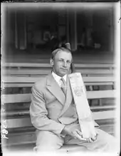 Man in double breasted suit, hair parted down the middle, sitting on a long bench in a sports stadium, posing with a cricket bat, held vertical and supported on his thigh.