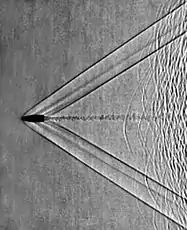 Shadowgram of shock waves from a supersonic bullet fired from a rifle.  The shadowgraph optical technique reveals that the bullet is moving at a Mach number of about 1.9. Left- and right-running bow waves and tail waves stream back from the bullet, and its turbulent wake is also visible. To the right of the frame are seen unburned powder particles from the cartridge, themselves traveling slightly subsonic so that each of them produces a curved bow shock wave.