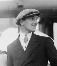 A photo of a clean-shaven Caucasoid man in a suit and flat cap; he is bodily facing the camera, though looking to his left.