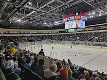 A 3/4 view of an ice hockey rink. The scoreboard says HEB Center and has a close-up shot of a hockey goalie. There are two teams on the ice, one in green and white and the other in maroon.