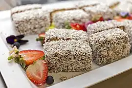 Lamingtons on a plate.  Each rectangular portion is completely coated on all sides by shredded coconut