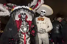 A man and woman in elaborate mariachi-style dress with skull paint